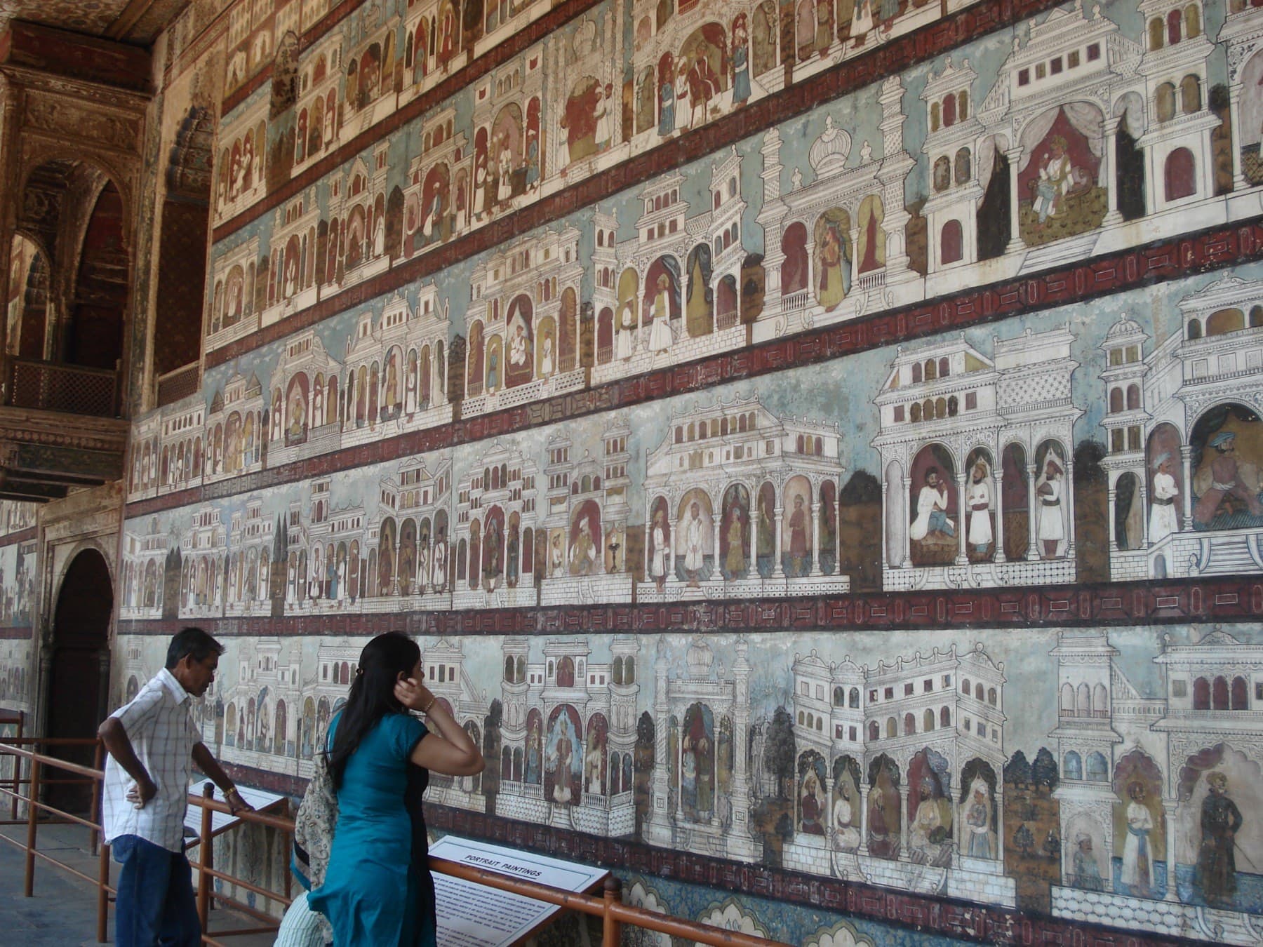 The east wall of Daria Daulat Bagh: five horizontal registers of painted palace scenes and seated rulers, with visitors standing before them.