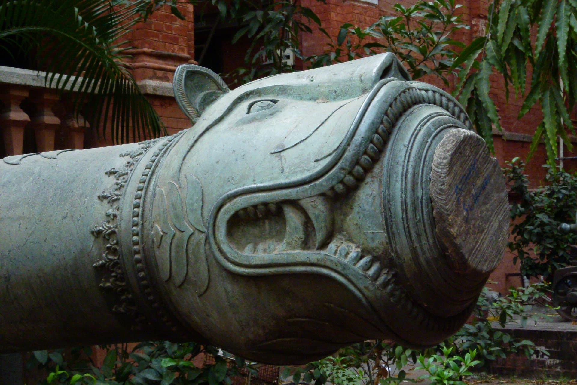 Close view of a bronze cannon muzzle cast as a snarling tiger's head, jaws open around the bore.