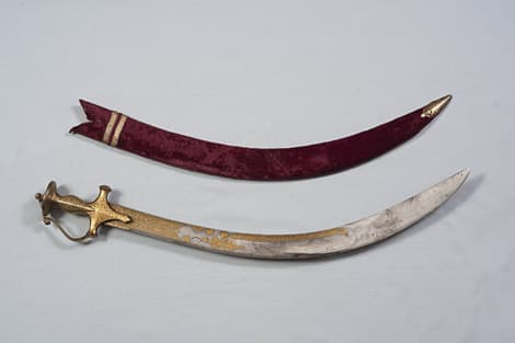 A curved sword of Tipu Sultan with a gold-damascened hilt and an inscribed blade, photographed against a dark background.