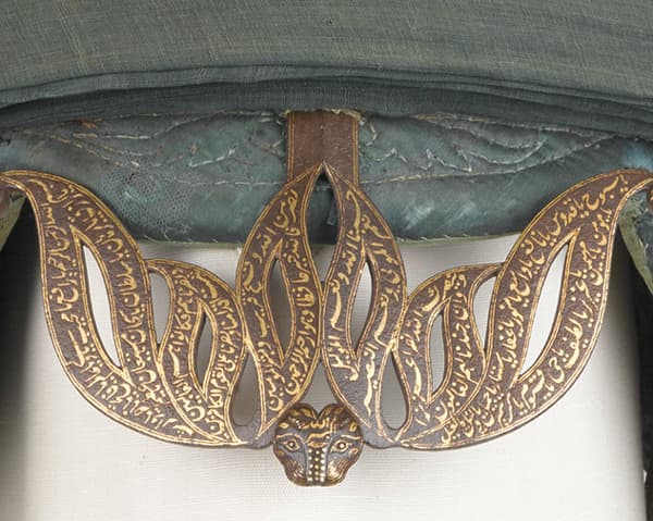 Close view of the war turban's guard: pierced metalwork in the form of tiger stripes filled with gold Arabic calligraphy, converging on a small tiger's head.
