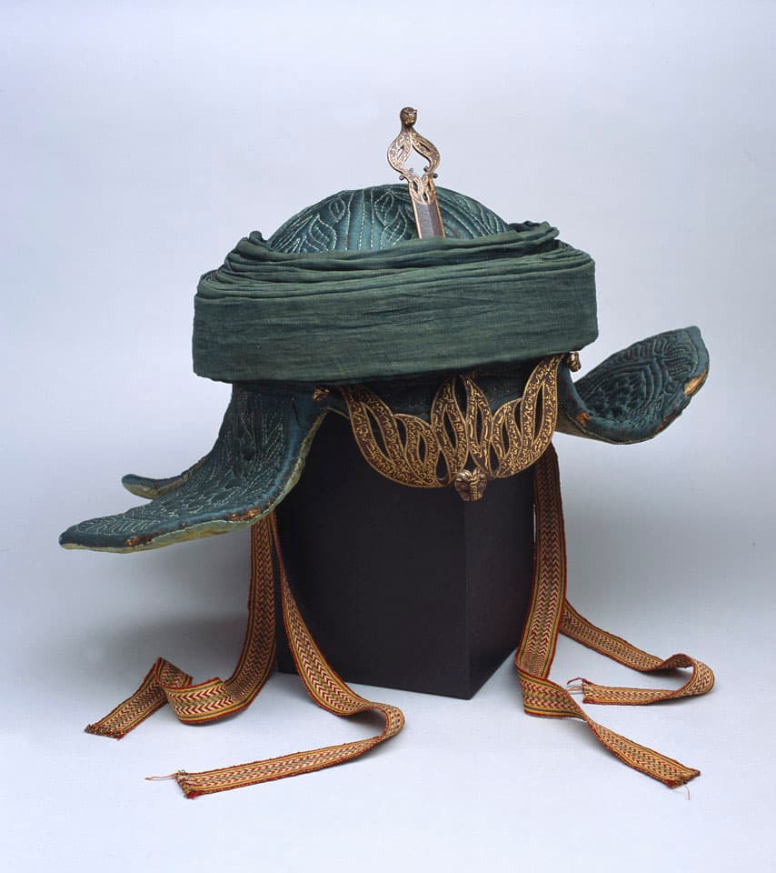 Tipu Sultan's war turban seen from the front: a quilted green helmet with a wound pagri, a gold openwork guard hanging at the front, and long woven ties trailing down over the display mount.